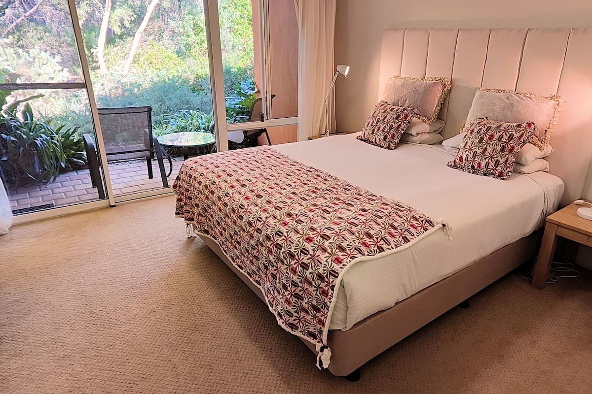 Une chambre avec un grand lit, une table de chevet, une chaise et une vue sur un jardin, visible à travers une porte vitrée.