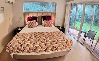 Una cama grande con cubrecama floral, dos mesitas de noche, una ventana con persianas y vista a un jardín a través de un ventanal.