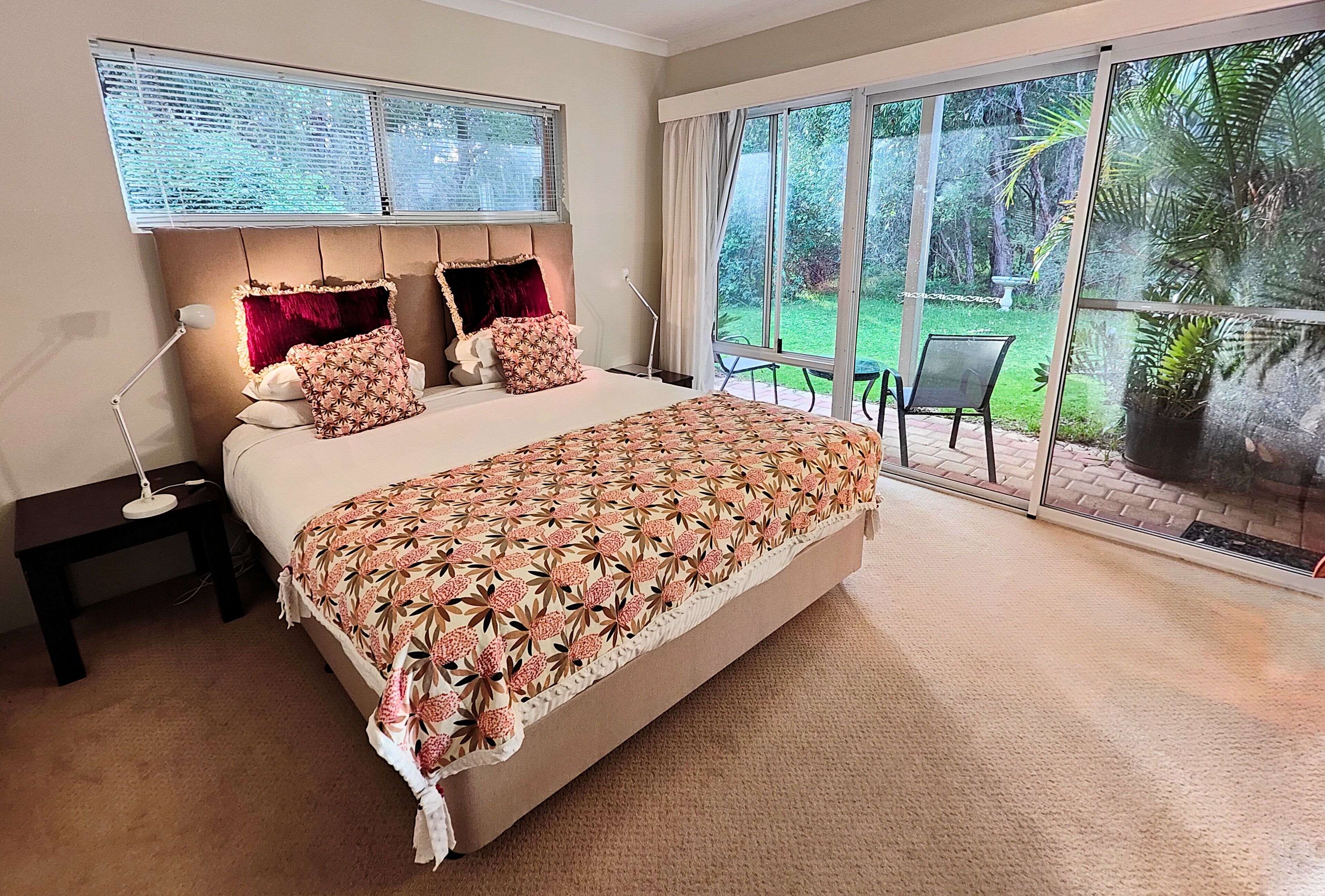 A bedroom with a large bed, a nightstand, a lamp, and a view of a garden through a sliding glass door.