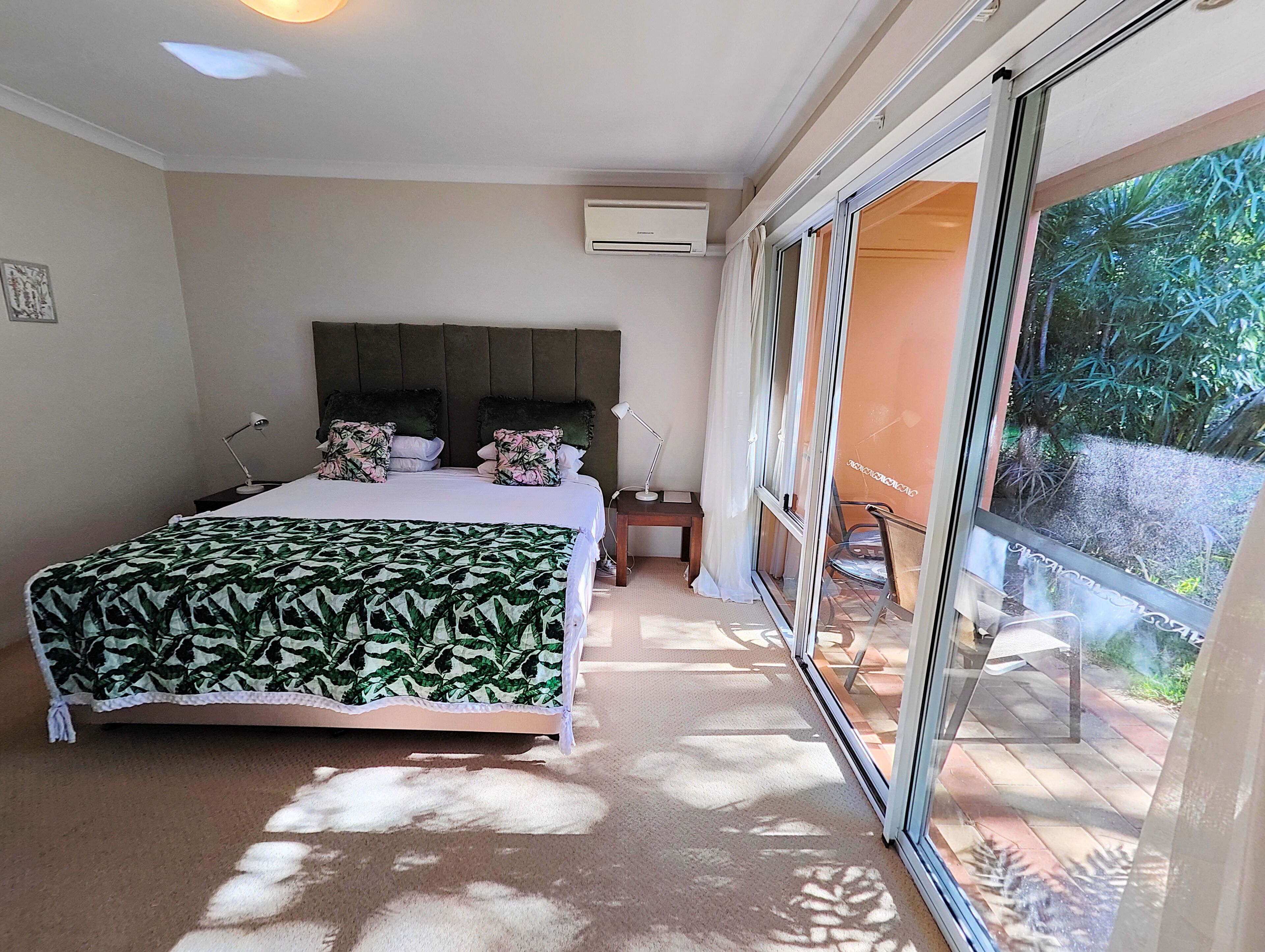 Un dormitorio con cama, mesita de noche, lámpara, ventana con vistas a zonas verdes y un equipo de aire acondicionado.