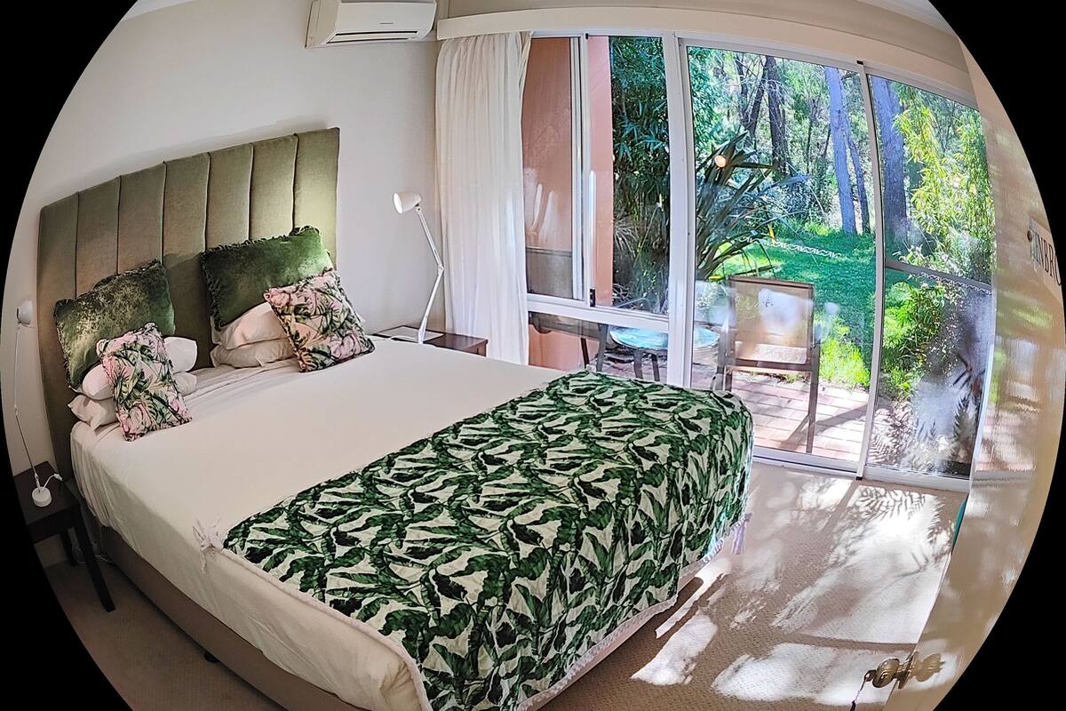 Une chambre à coucher avec un grand lit, une table de chevet, une lampe et une fenêtre donnant sur un jardin.