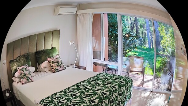A bedroom with a large bed, a nightstand, a lamp, and a window with a view of a garden.