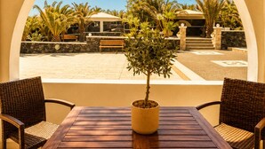 A wooden table with a potted plant in the center, flanked by wicker chairs, under a large arched window overlooking a patio area with a bench and palm trees. - Villa Danezis (Santorini)