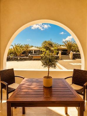 A wooden table with a potted plant in the center, flanked by wicker chairs, under a large arched window overlooking a patio area with a bench and palm trees. - Villa Danezis (Santorini)