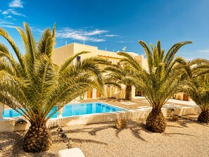 A pool area with palm trees, a clear blue sky, and a building with a flat roof. - Villa Danezis (Santorini)