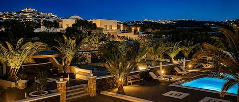 Área de piscina en un resort con sillones de descanso y palmeras, rodeada por un muro de piedra y vista a los edificios del resort.