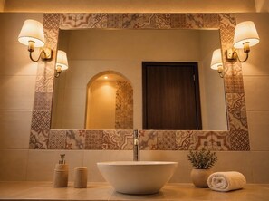 A bathroom with a large mirror, two wall-mounted lamps, a vessel sink, a towel, and a small potted plant. - Villa Danezis (Santorini)