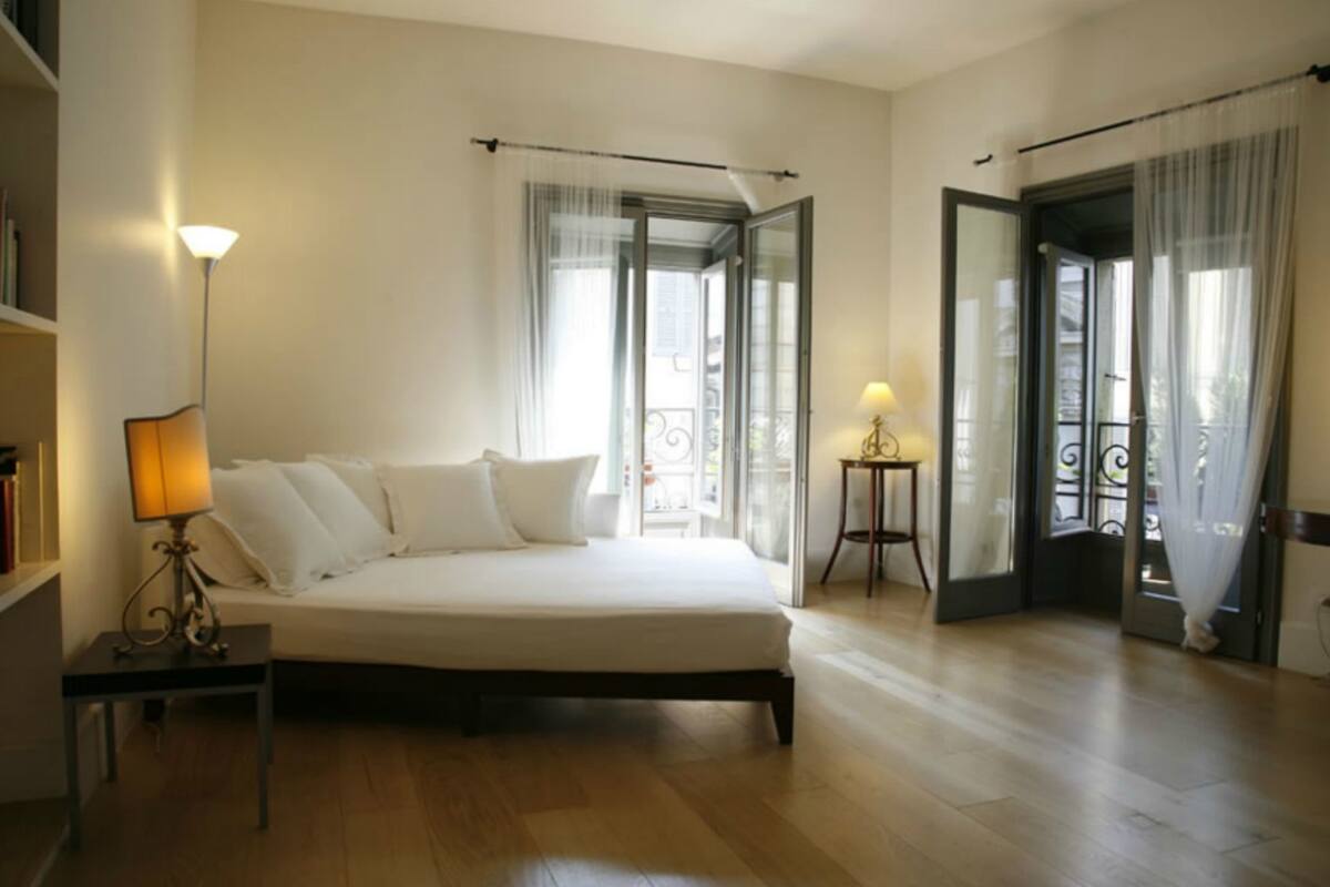 A bedroom with a bed, a bedside table with a lamp, a bookshelf, and a balcony with a railing.
