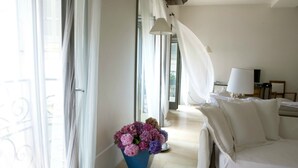 A hotel room with a bed, a desk with a lamp, a window with sheer curtains, and a potted plant with flowers. - Milanosuites (Milan)