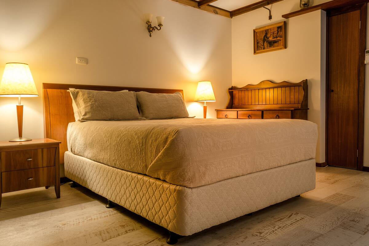 A neatly made bed with a quilted cover, two bedside lamps, a wooden nightstand with drawers, a wooden headboard, and a painting on the wall.