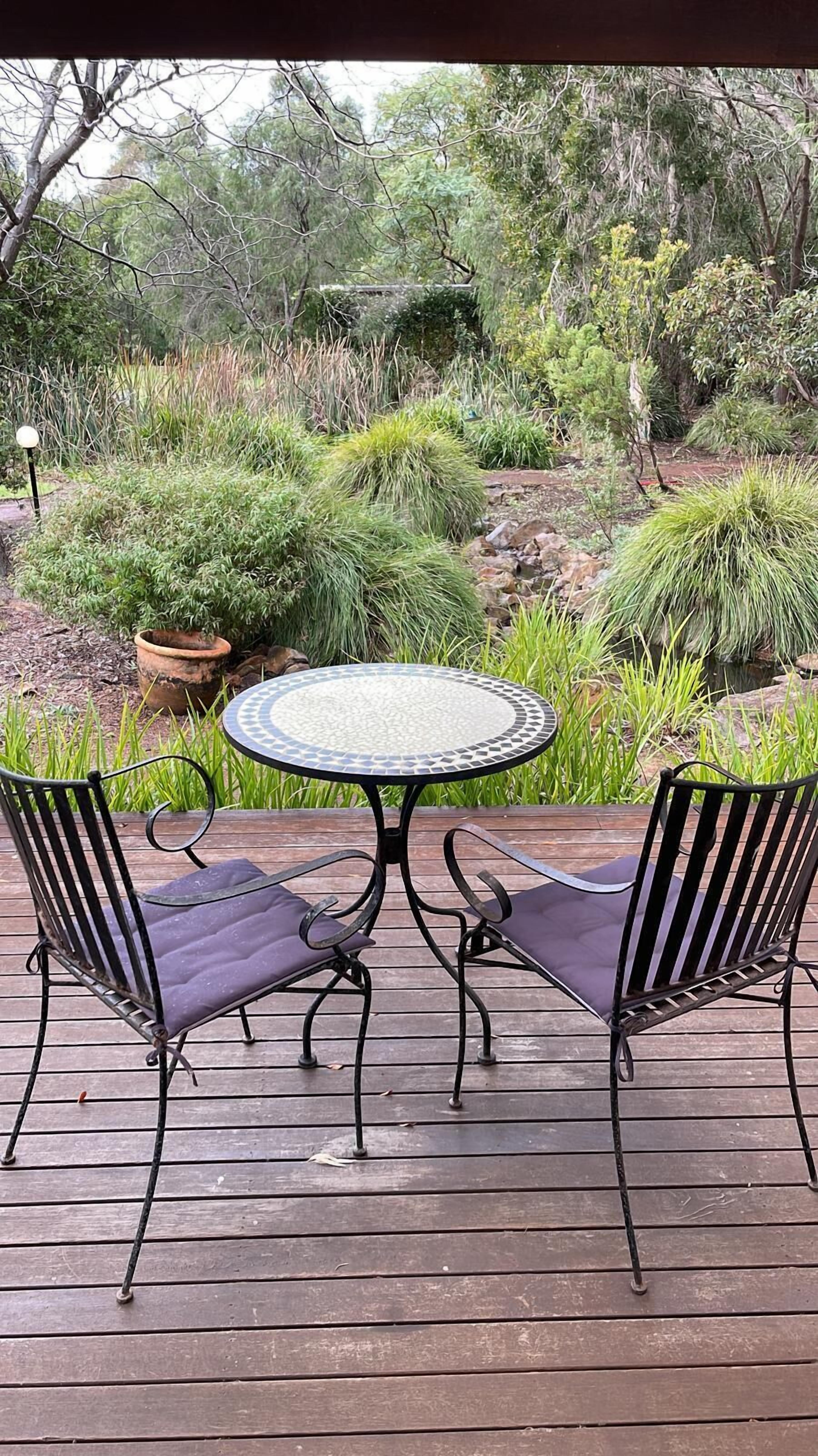 A round table with a mosaic top surrounded by two cushioned chairs with metal frames. The table is set on a wooden deck with a natural backdrop of various plants and trees.
