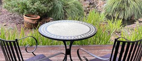 A round table with a mosaic top surrounded by two cushioned chairs with metal frames. The table is set on a wooden deck with a natural backdrop of various plants and trees.