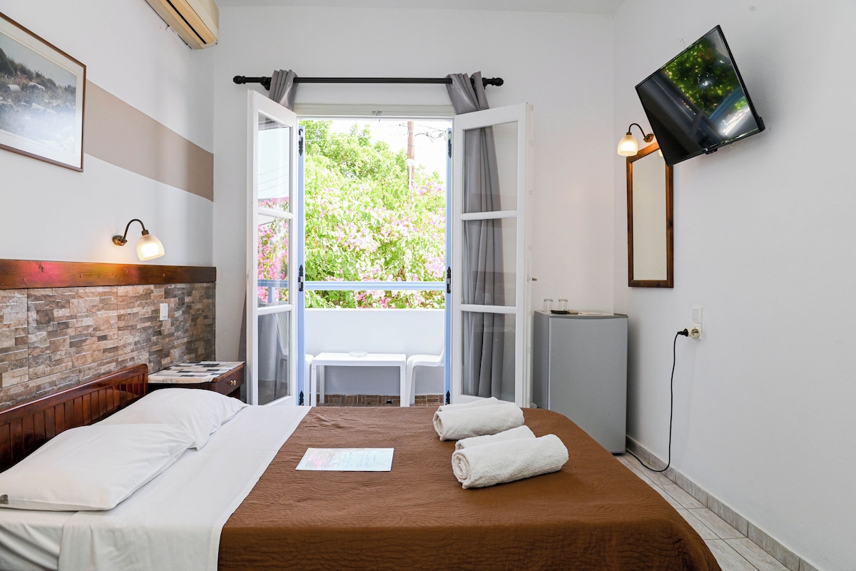 A hotel room with a bed, a desk, a chair, a TV, and a balcony with a view of greenery.