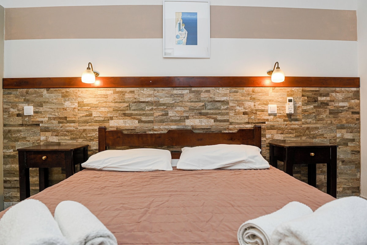 A hotel room with a stone wall, wooden headboard, two bedside tables, and a bed with white pillows and a pink bedspread.