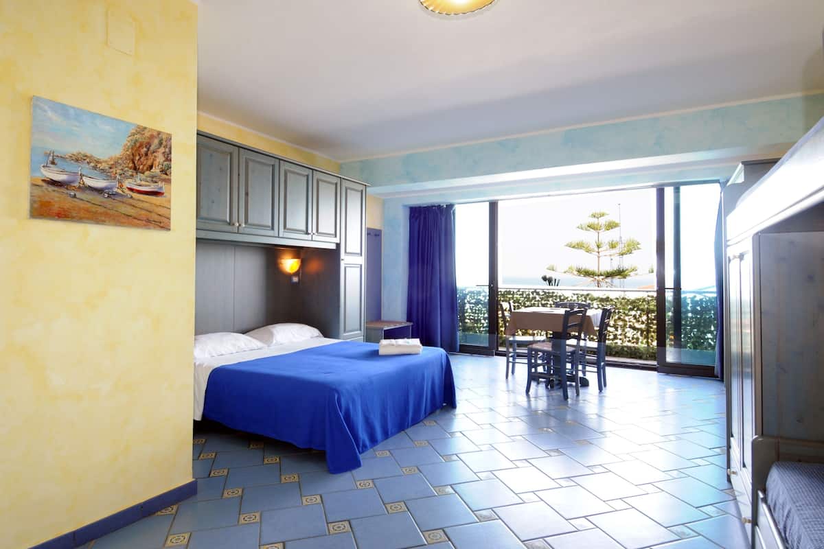 Ein Hotelzimmer mit Bett, Schreibtisch, Stuhl, einem Balkon mit Aussicht und einem Bild an der Wand.