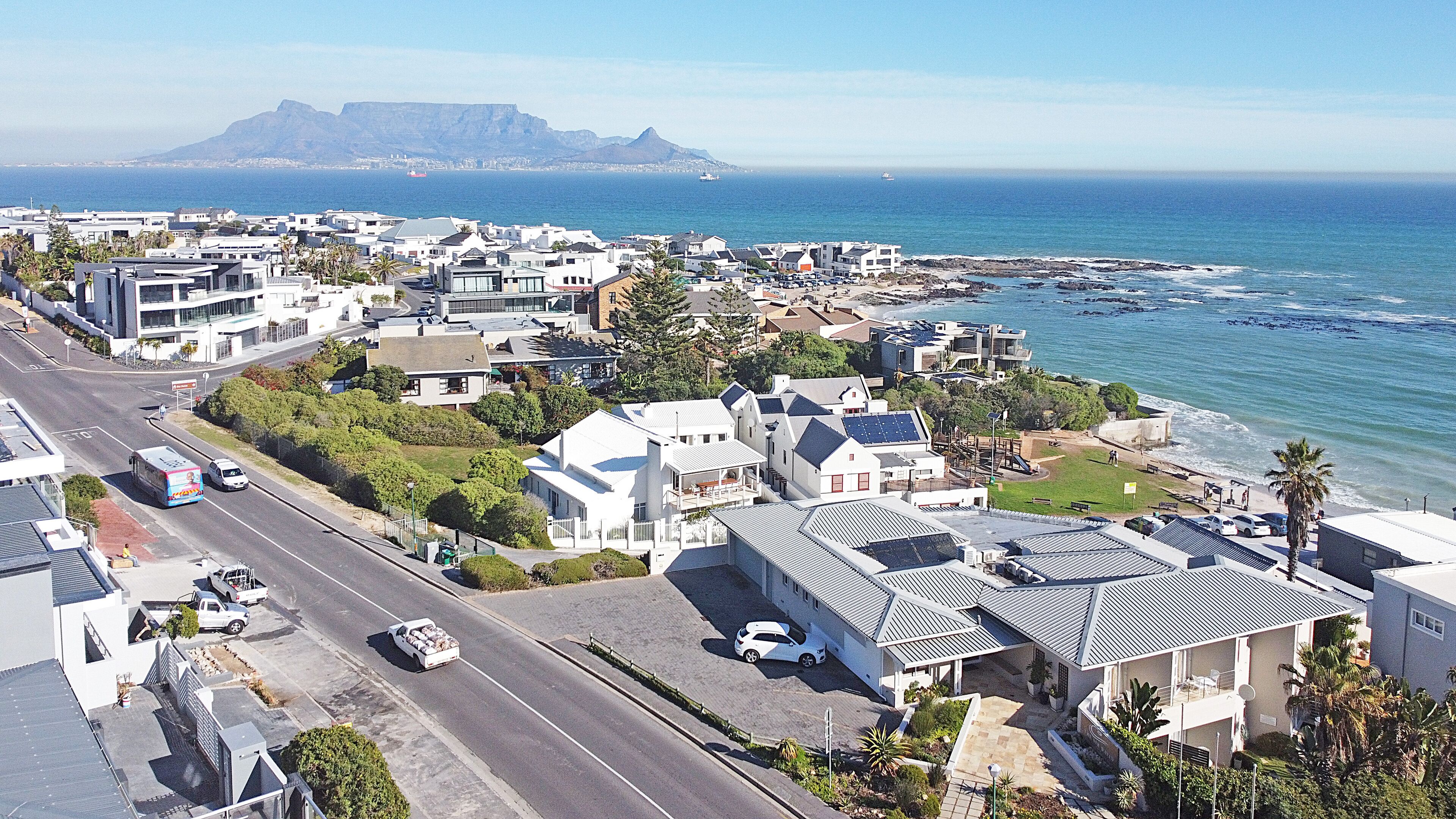 A coastal residential area with modern houses, a road, and a view of the ocean and mountains in the background.