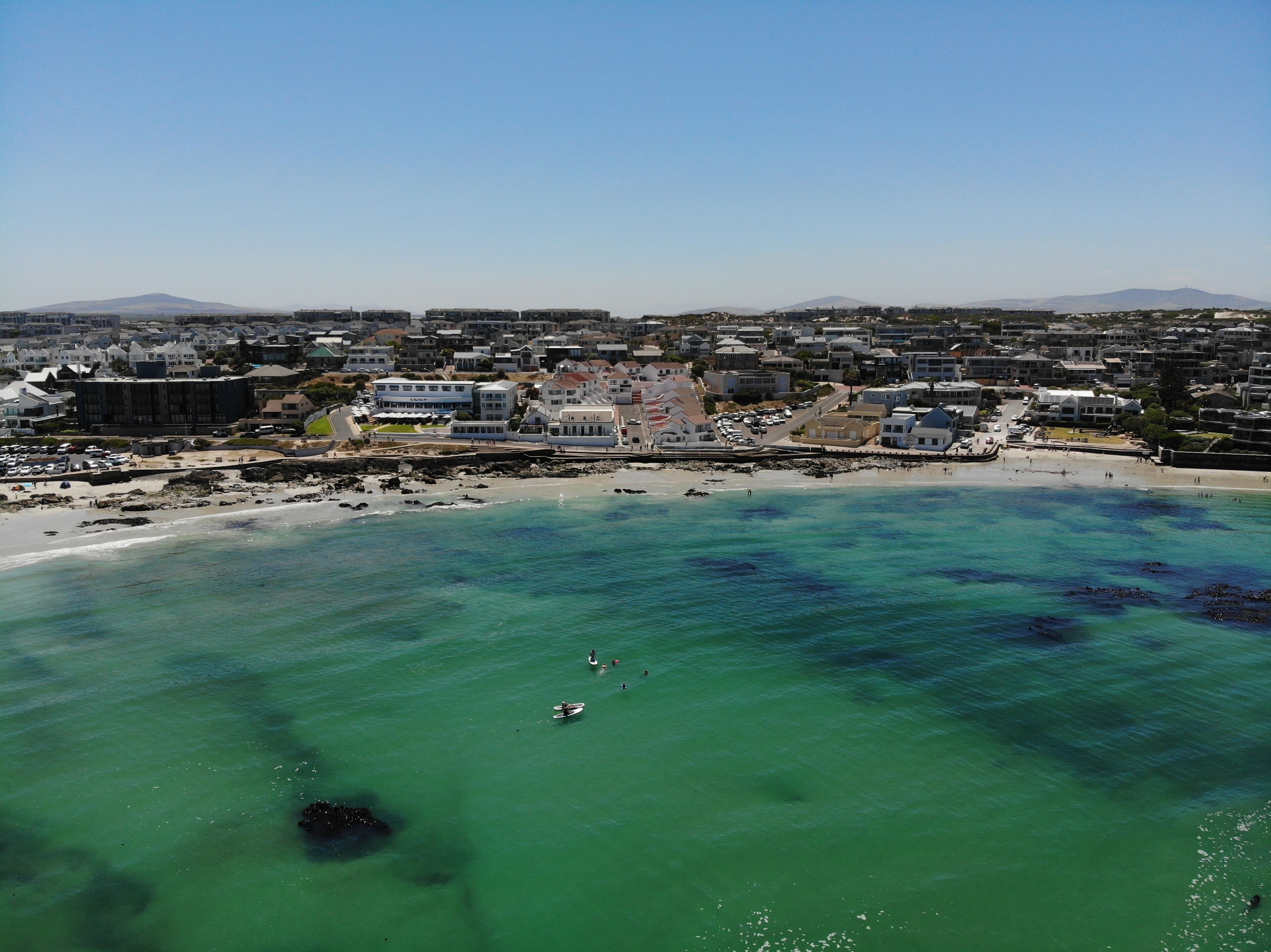 An aerial view of a coastal town with a beach, buildings, and a mountain in the background.