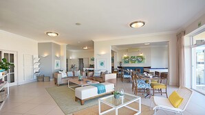 Een ruime woonkamer met een bank, salontafel en eettafel. Er staan rieten stoelen en een witte fauteuil. De kamer heeft een groot raam en een deur die leidt naar een andere ruimte.
