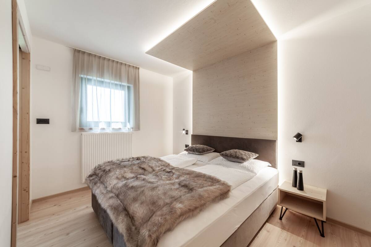 A modern bedroom with a large bed, a wooden headboard, a window with curtains, and a small bedside table with two bottles.