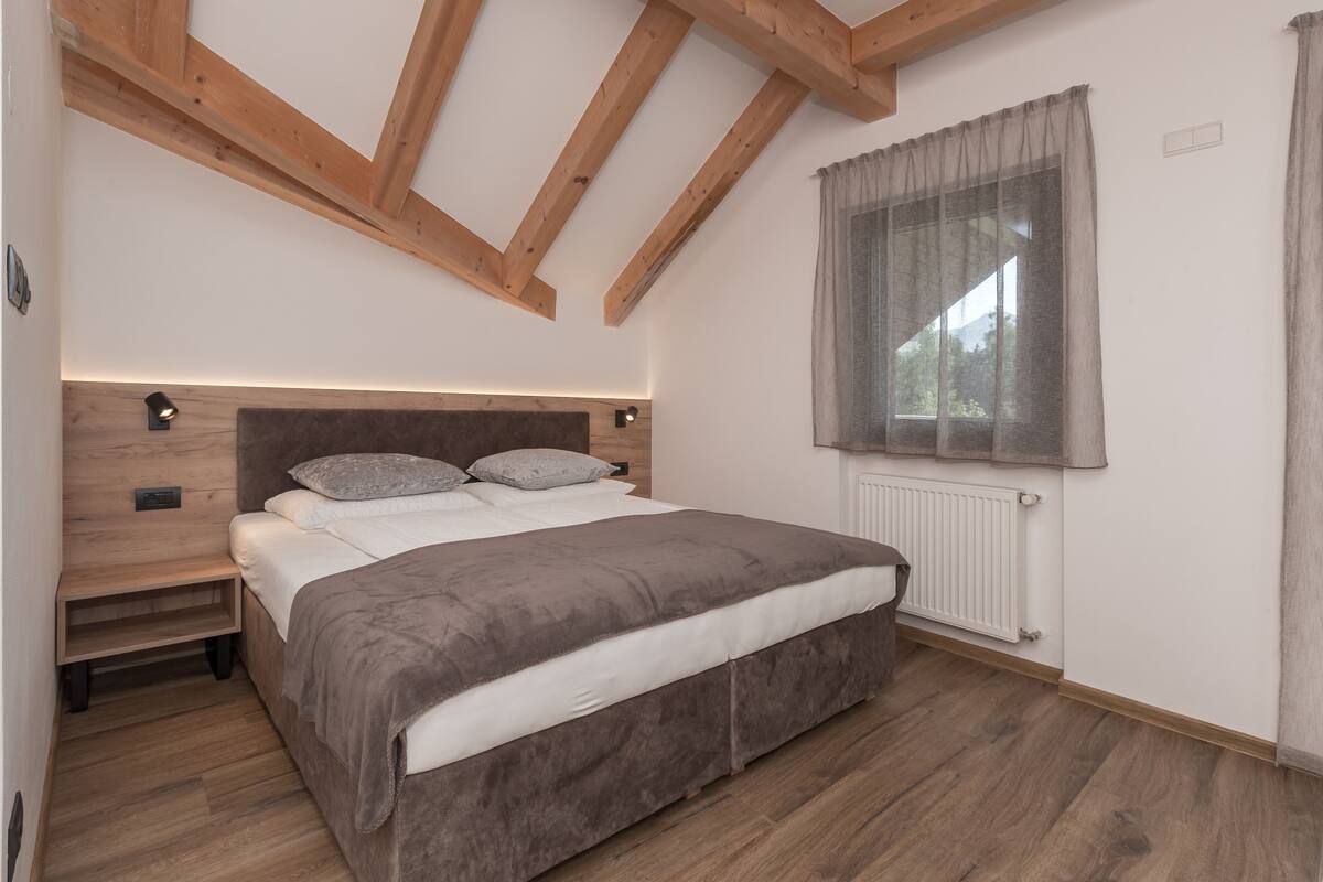 A bedroom with a wooden ceiling, a bed with a grey duvet, a bedside table, and a window with curtains.
