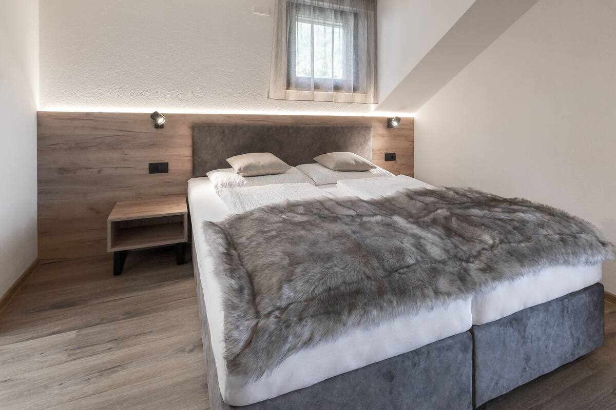 A modern bedroom with a wooden headboard, a large bed with a grey fur throw, and a small bedside table.