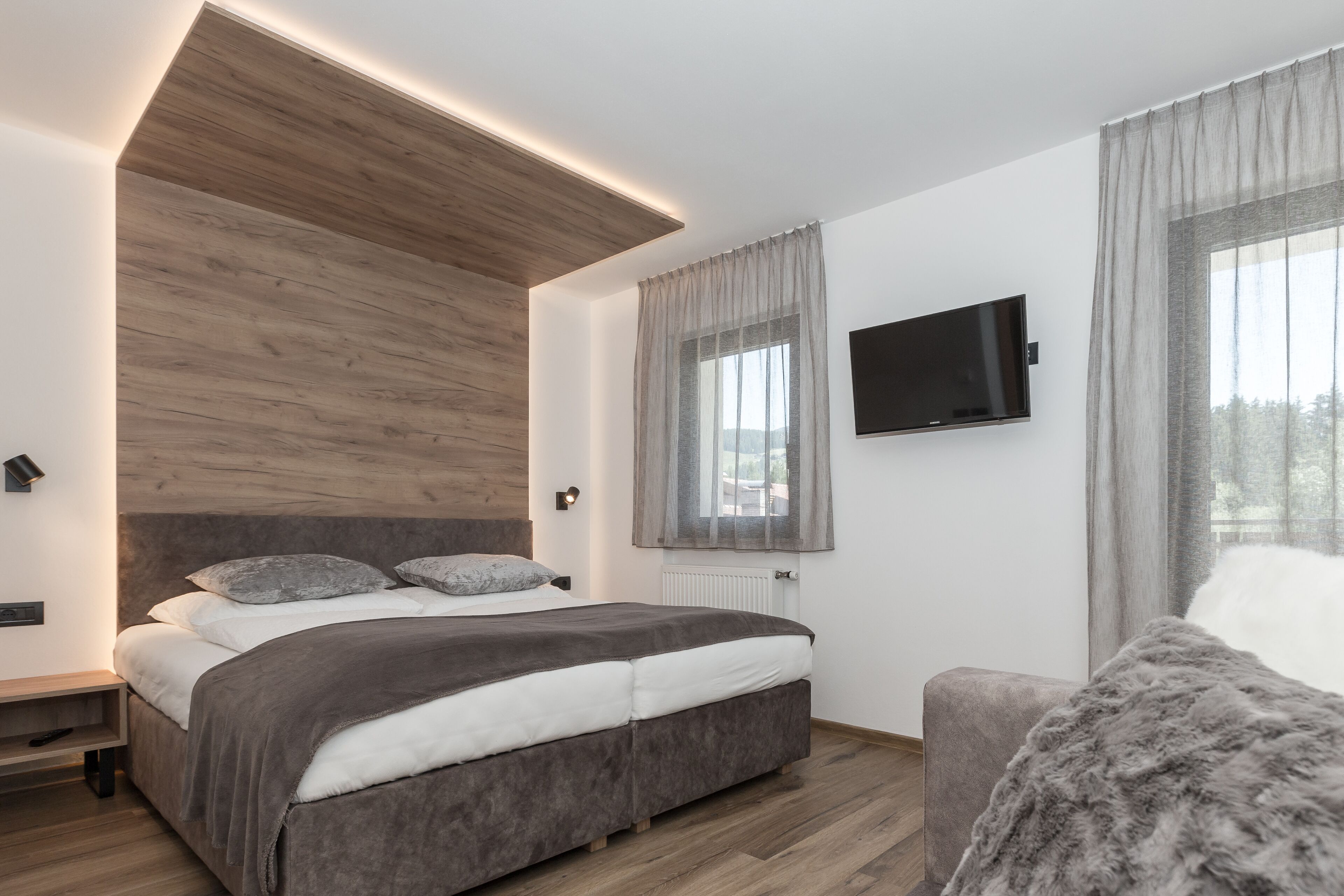 A modern bedroom with a bed, a TV mounted on the wall, and a window with curtains.