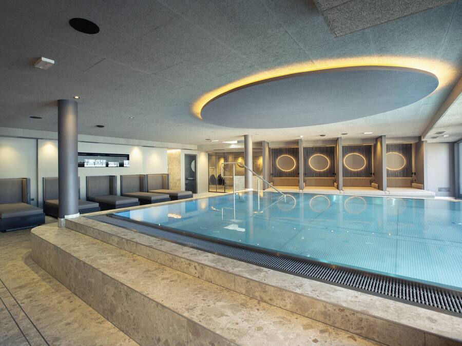 A modern indoor swimming pool with a geometric ceiling design, seating areas, and a clean, contemporary aesthetic.