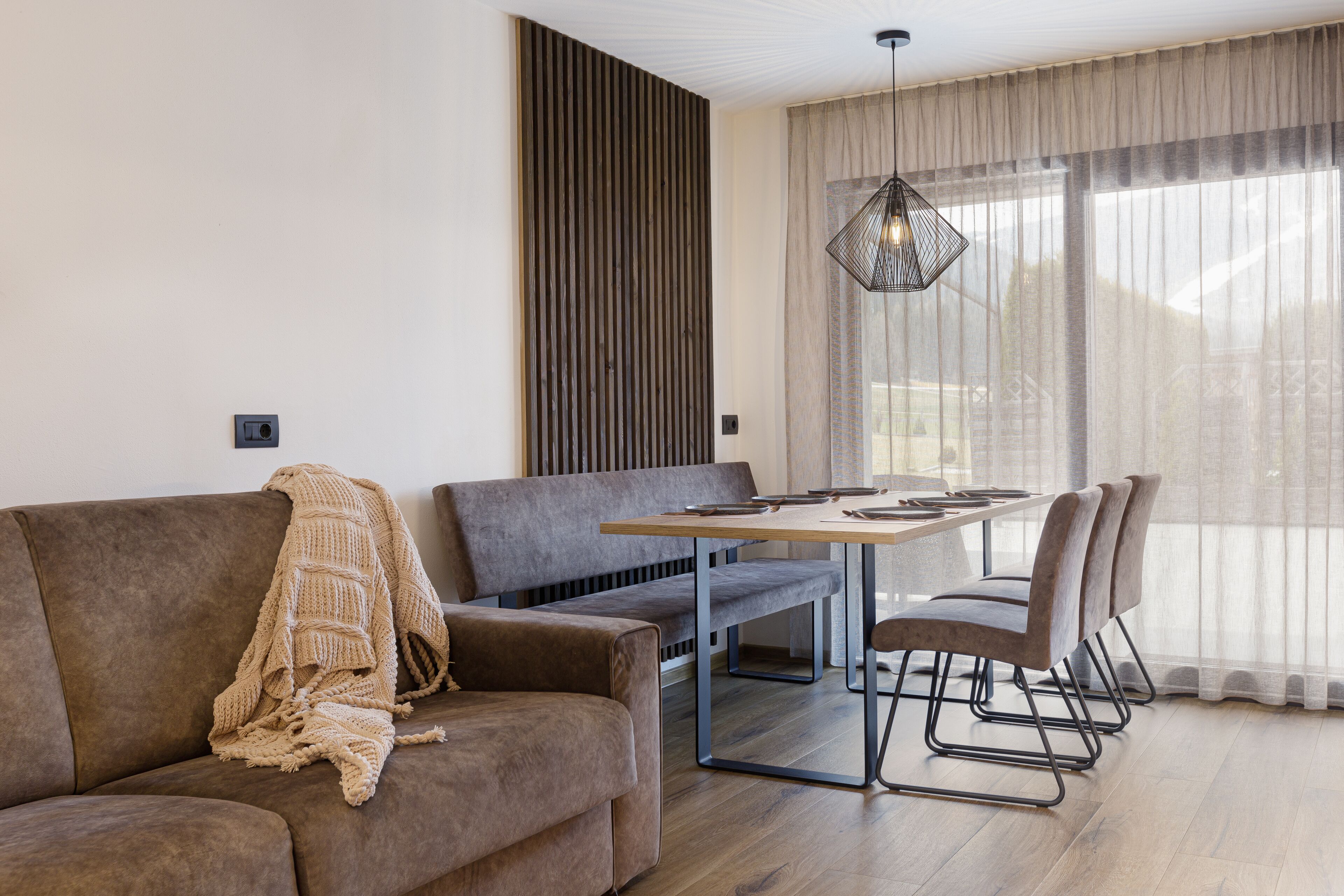 En moderne stue med sofa, bænk, spisebord med stole, pendel og et stort vindue med lette gardiner.
