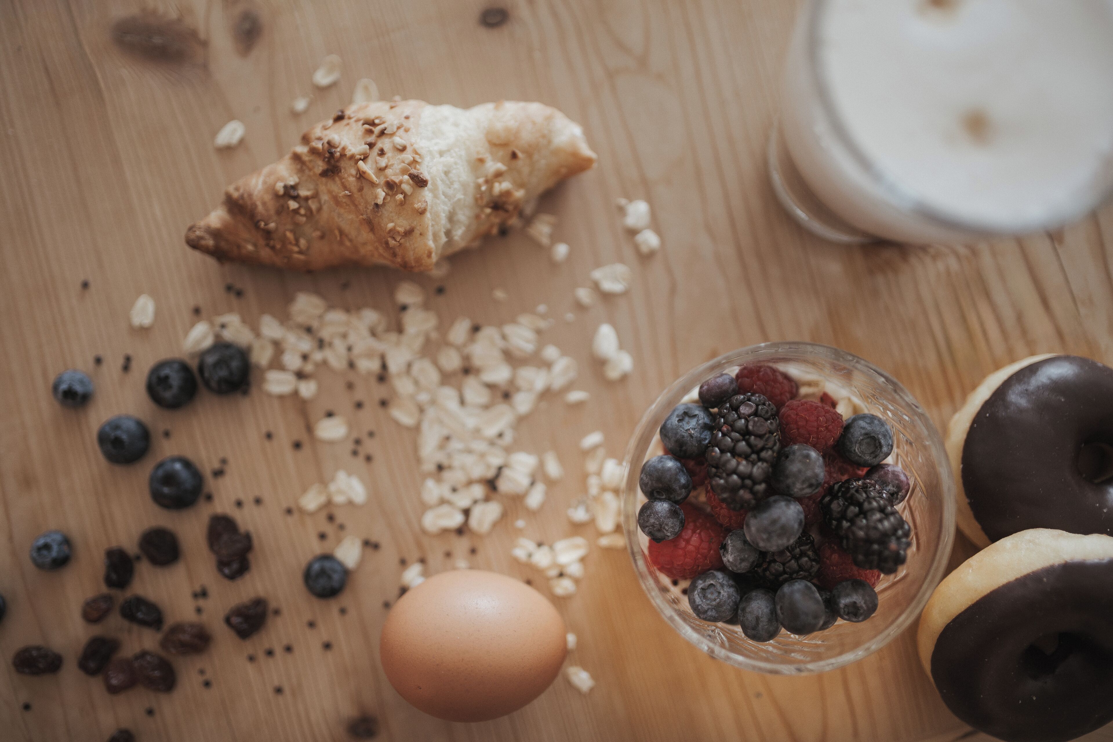 Une table en bois avec un croissant, un bol en verre rempli de fruits rouges variés, un beignet au chocolat et un verre de lait.