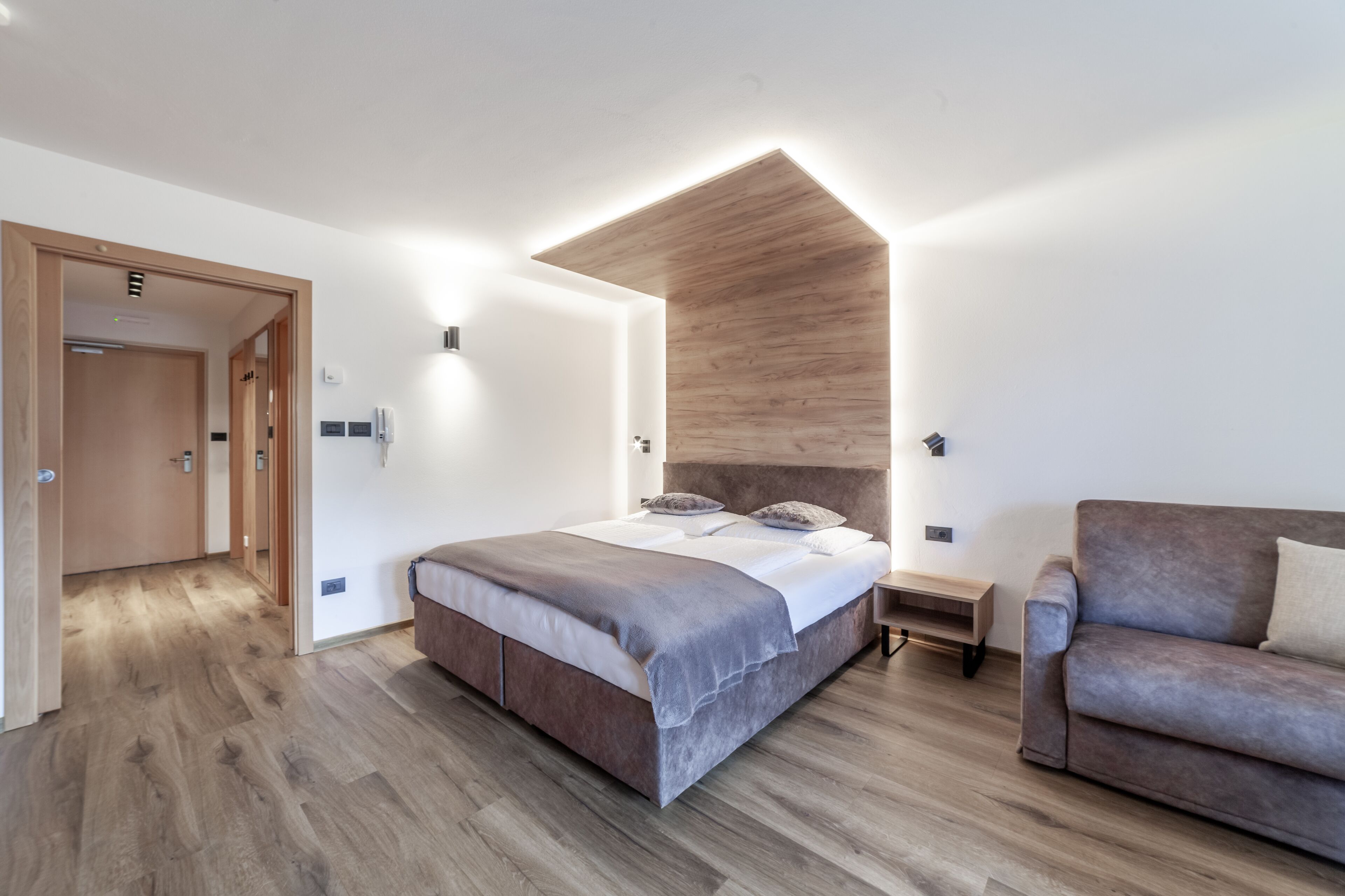 Un dormitorio moderno con una cama grande, cabecera de madera, suelo de madera, un sofá gris y una mesita de noche pequeña.