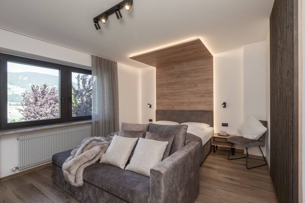 A modern bedroom with a large window, a sofa, a bed, and a bedside table.