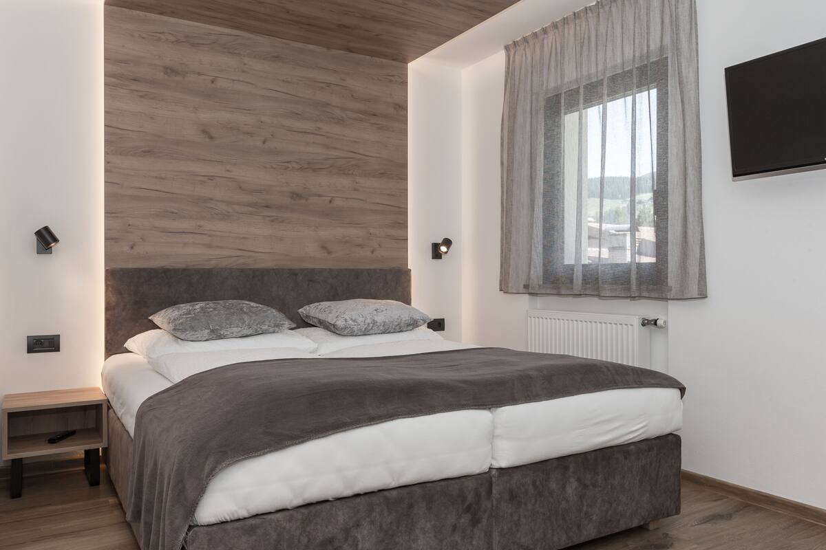 A modern bedroom with a wooden ceiling, a large bed with grey bedding, a bedside table, and a TV mounted on the wall.
