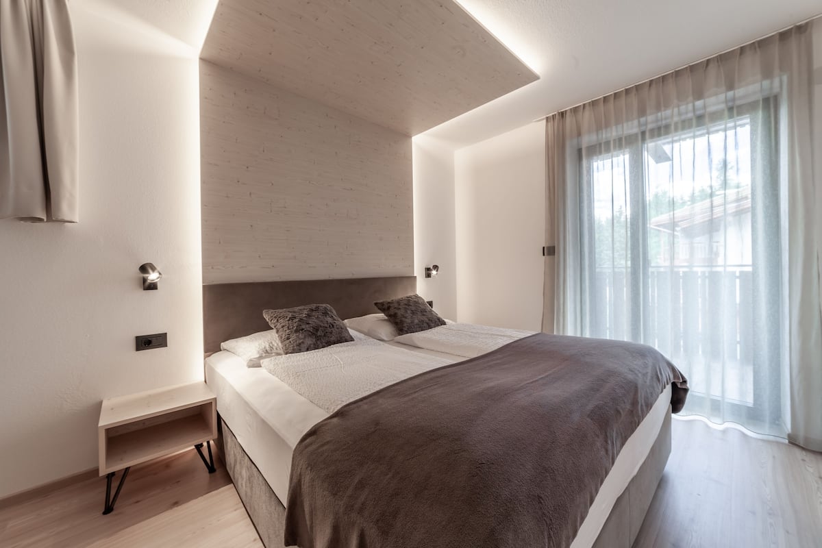A modern bedroom with a large bed, a bedside table, and a window with sheer curtains.