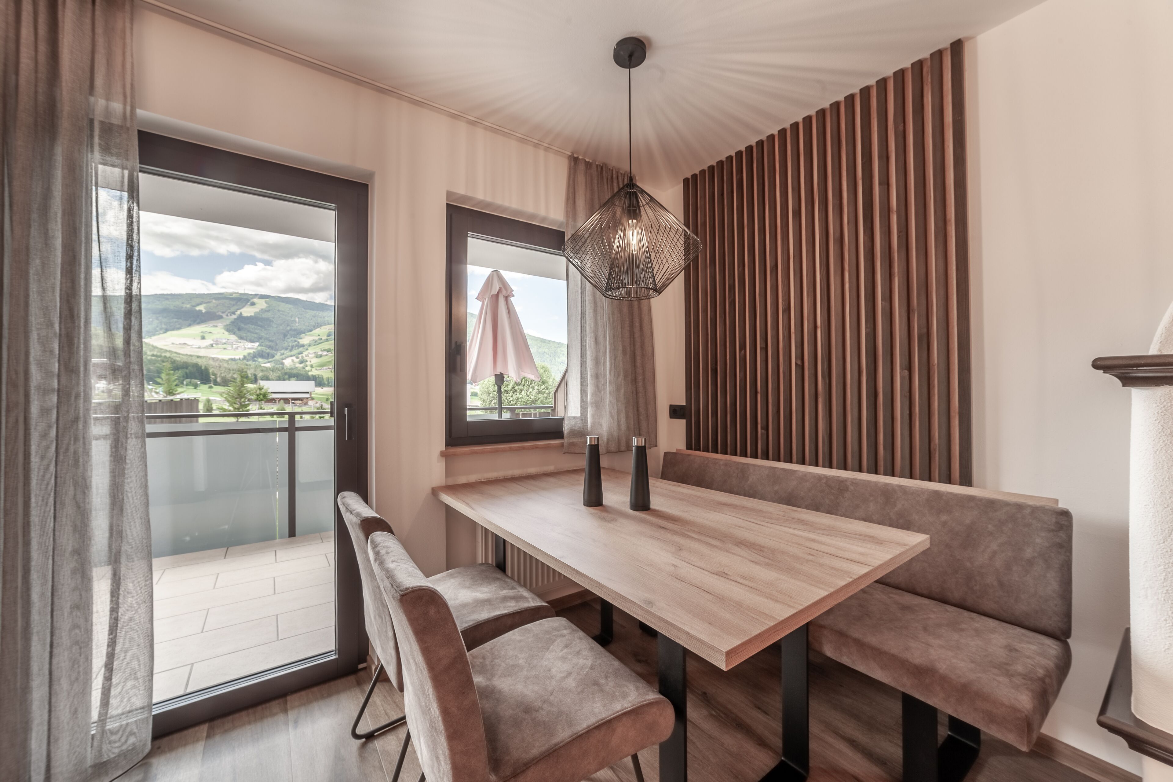A modern dining area with a wooden table and cushioned benches, a hanging light fixture, and a view of a balcony with a glass door.