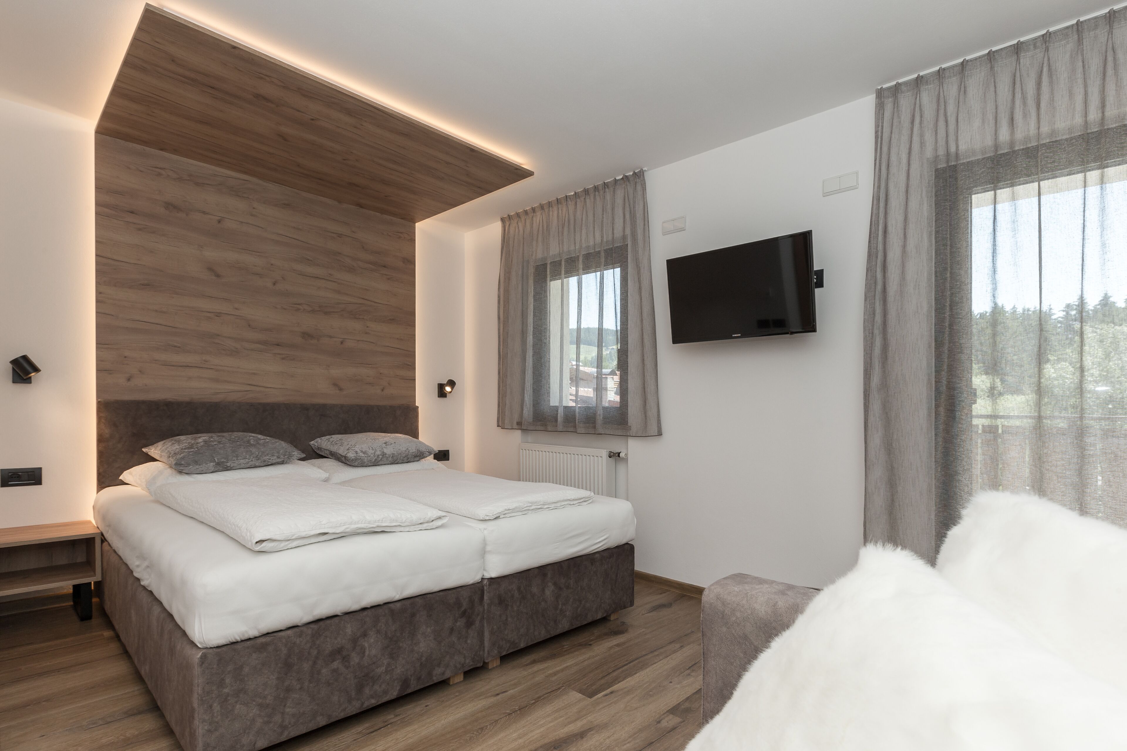 Un dormitorio moderno con cabecera de madera, cama con ropa de cama blanca, televisor montado en la pared y una ventana con cortinas.