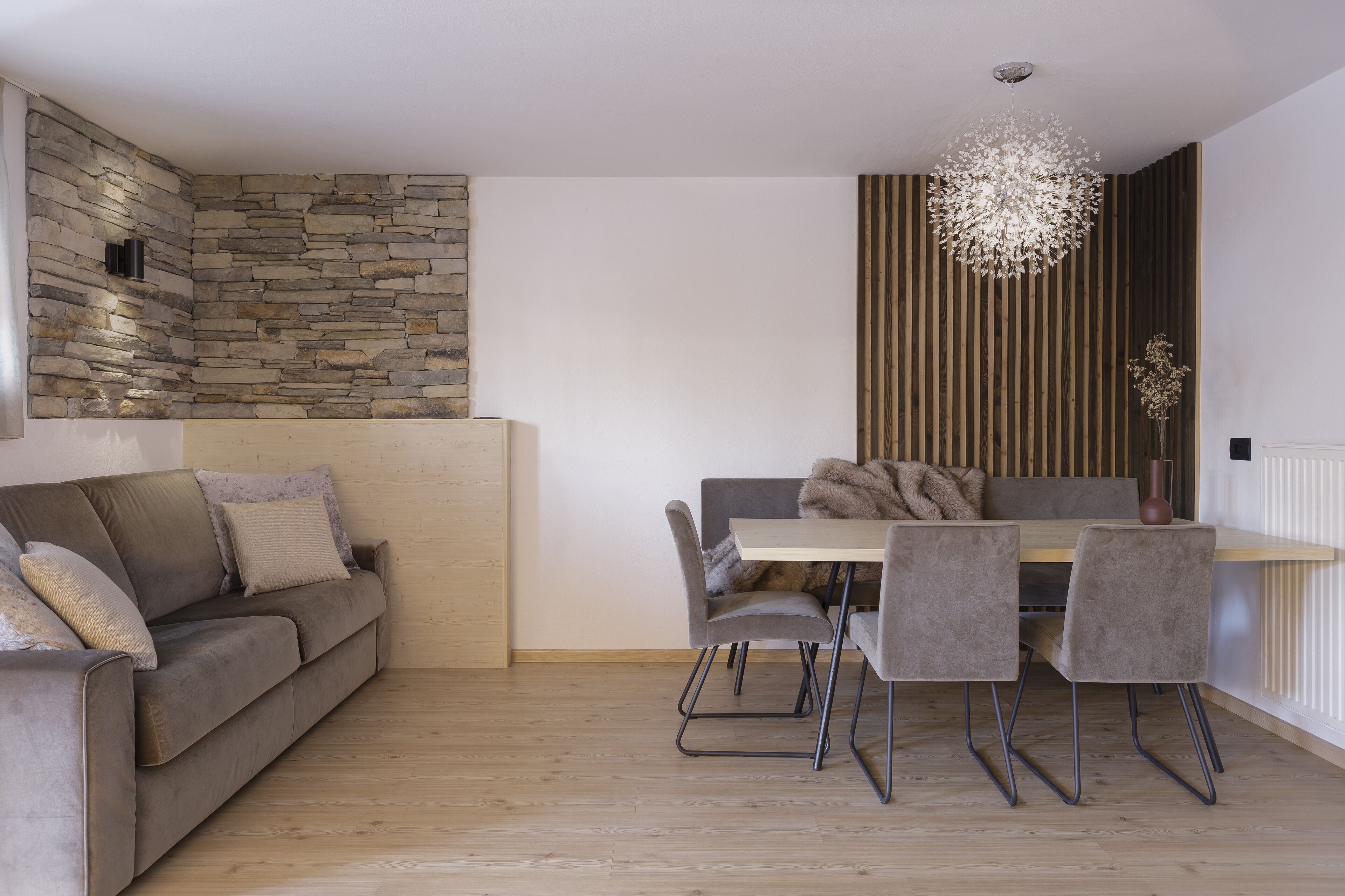 Una sala moderna con pared de piedra, pared de listones de madera, sofá gris, mesa de comedor con cuatro sillas y lámpara colgante.