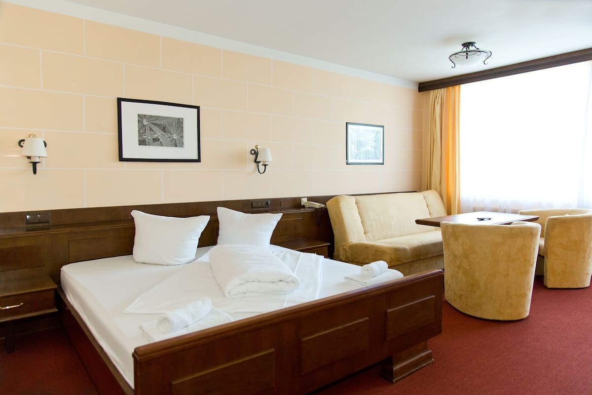 A hotel room with a large bed, a sofa, two armchairs, a small table, and a window with curtains.