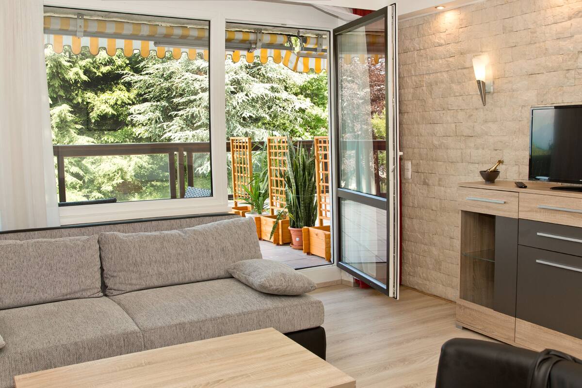 A modern living room with a sofa, a coffee table, and a TV on a cabinet. There is a sliding glass door leading to a balcony with potted plants.