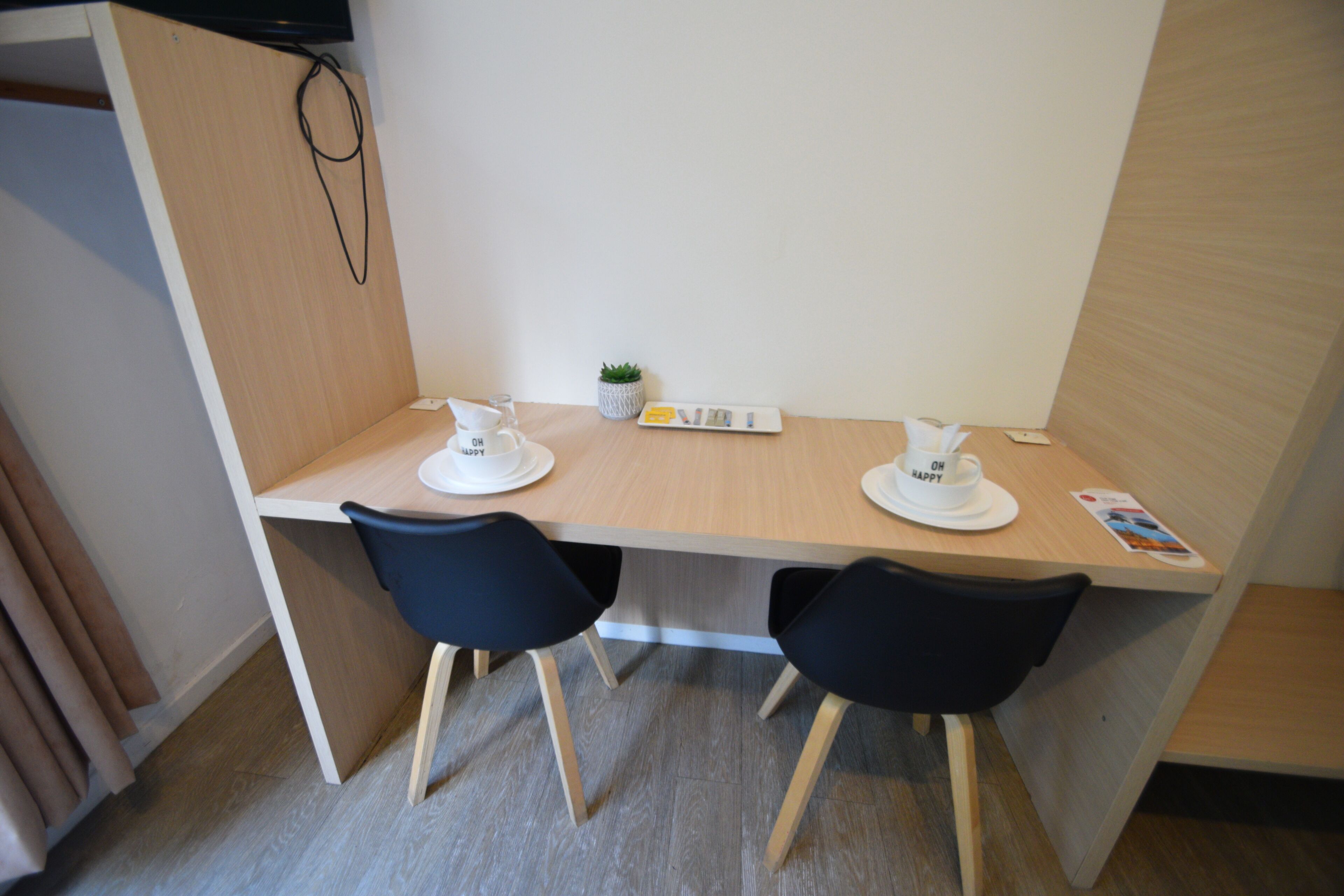 Une petite pièce moderne, avec un bureau en bois, deux chaises noires, une petite plante en pot et un téléviseur fixé au mur.