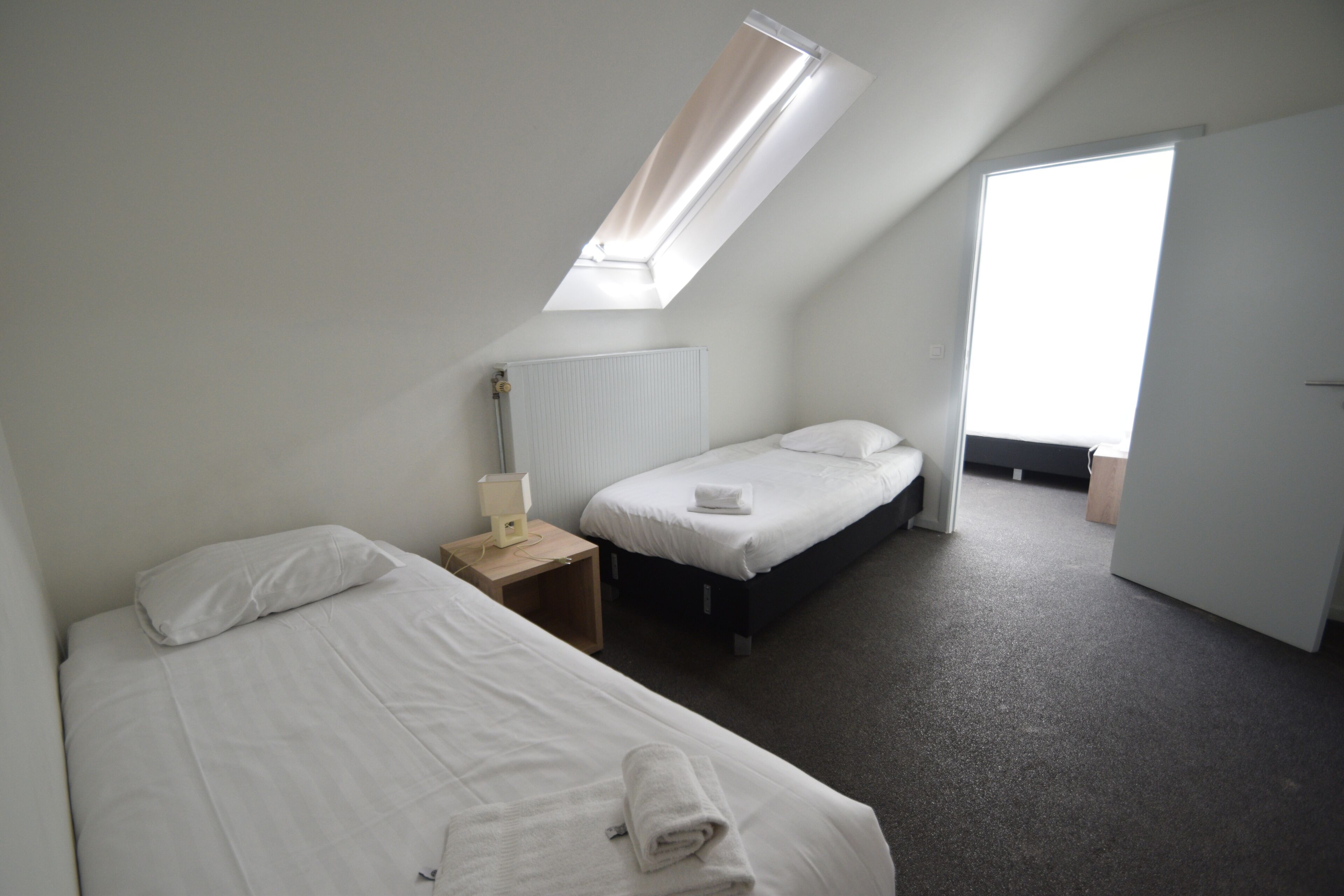 Une chambre avec deux lits, une lucarne, une table de chevet avec une lampe et une porte donnant sur une autre pièce.