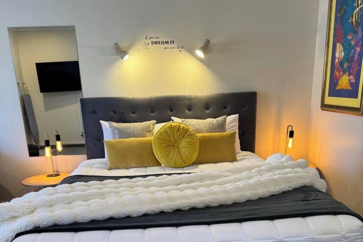 A bedroom with a tufted headboard, a large bed with white and black bedding, a yellow pillow, and a wall-mounted light fixture.