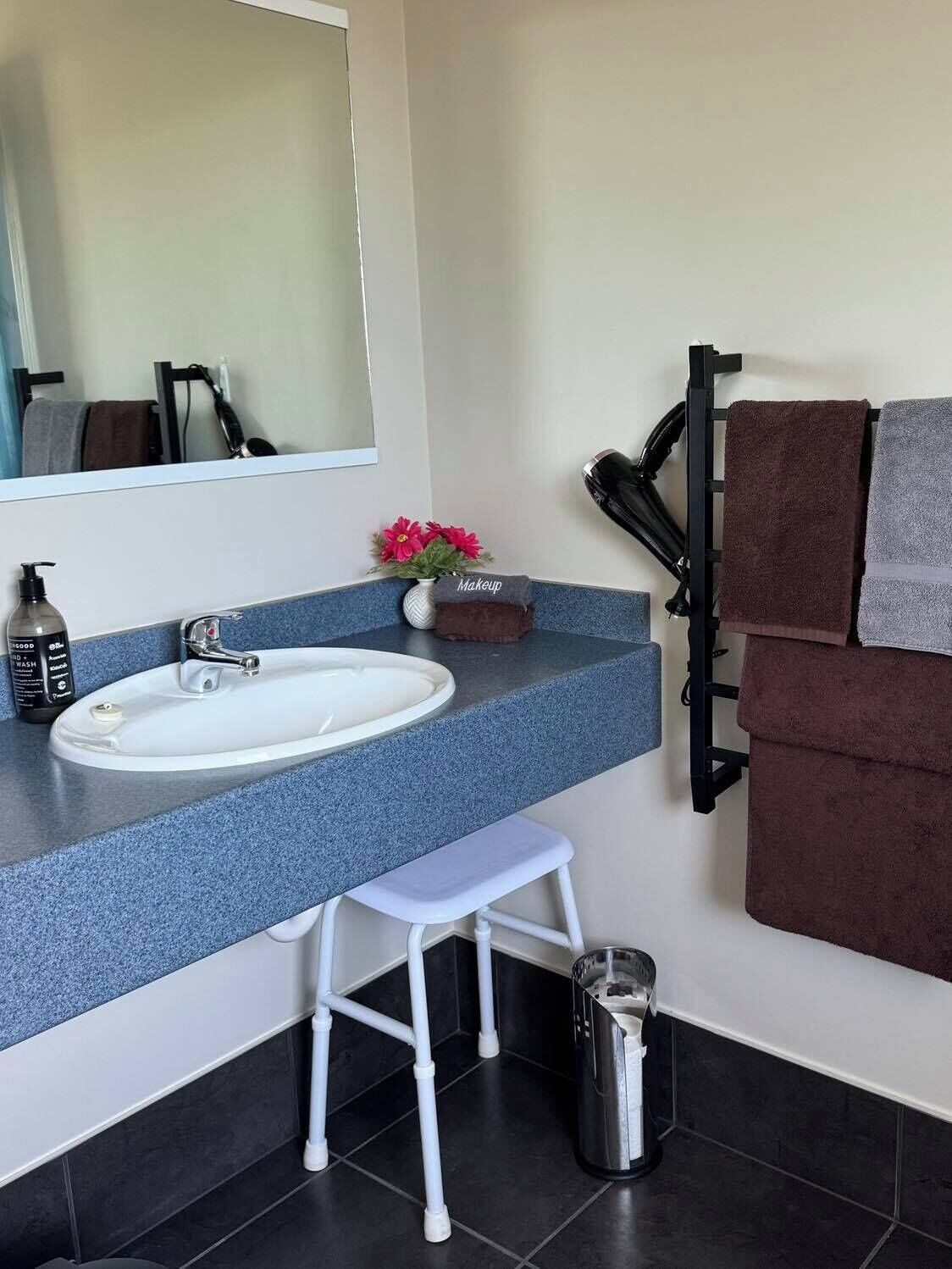A bathroom with a white sink, blue countertop, and a small white stool. There is a mirror above the sink, a towel rack with towels, and a trash can.