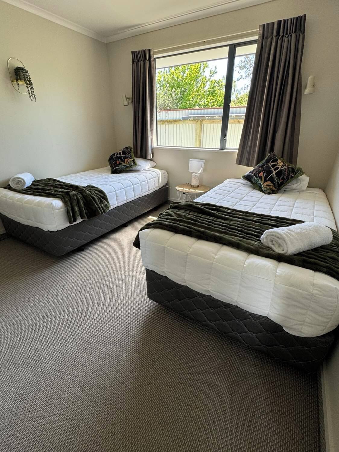 A hotel room with two beds, a small table with a lamp, and a window with curtains.