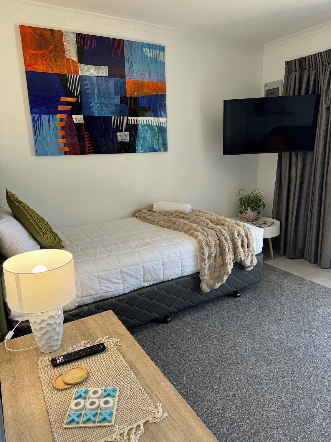 Habitación de hotel con cama, televisión, un cuadro y una mesa con lámpara y control remoto.