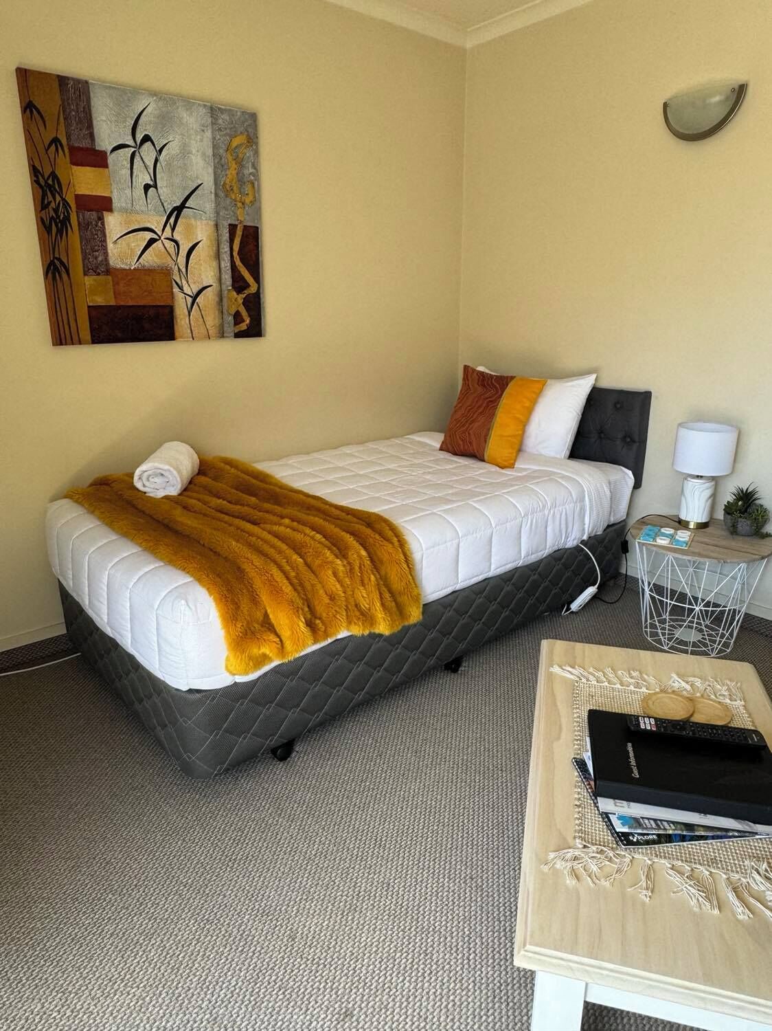 A single bed with a white duvet and a mustard-colored throw blanket, a bedside table with a lamp, and a wall-mounted light fixture.
