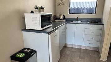 A compact kitchen with a microwave, sink, and cabinets. There's a small trash bin and a window with blinds.