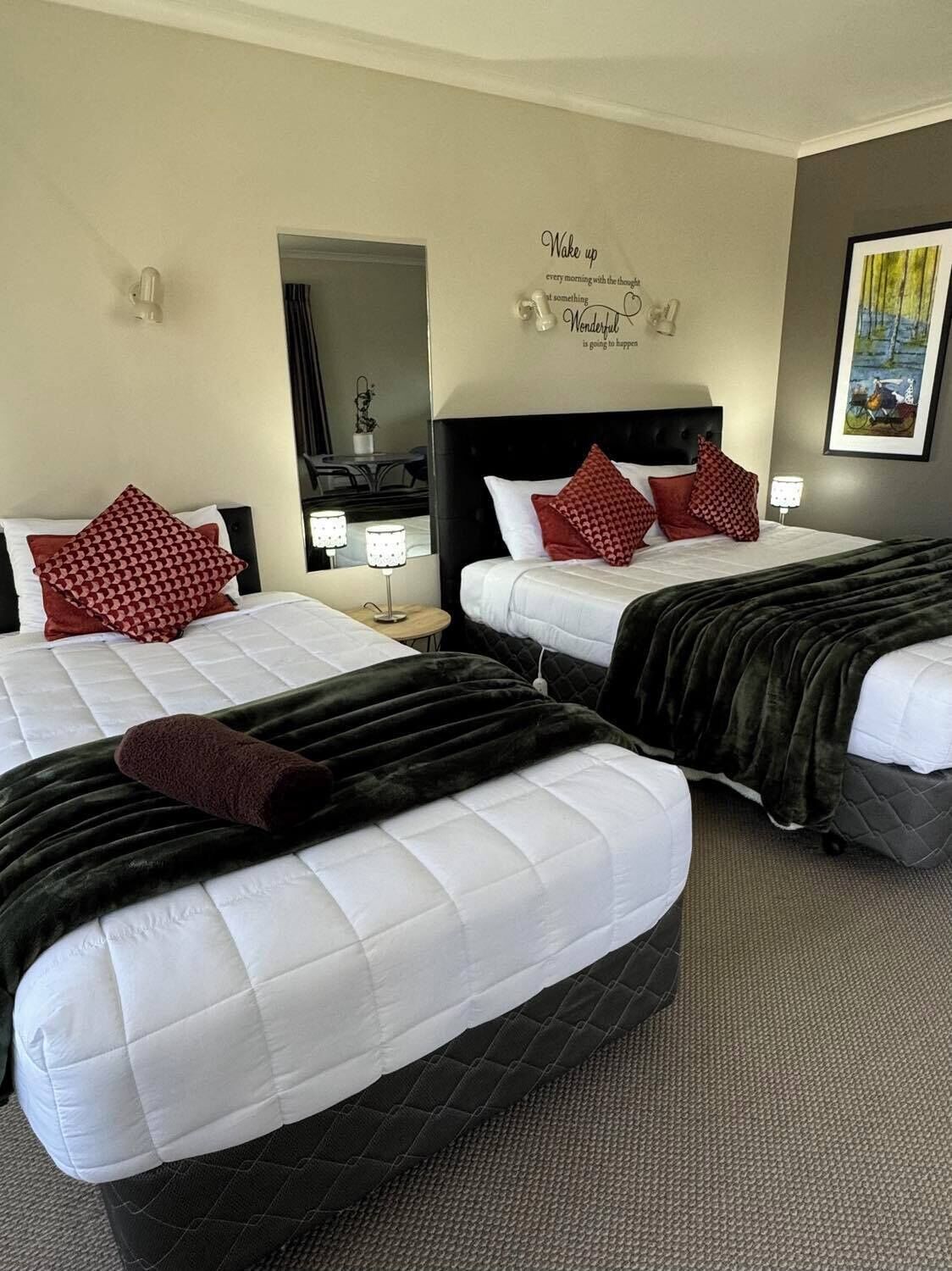 A hotel room with two beds, each with white bedding and red patterned pillows. There is a black headboard on one bed and a mirror above the bed. A motivational quote is displayed on the wall.