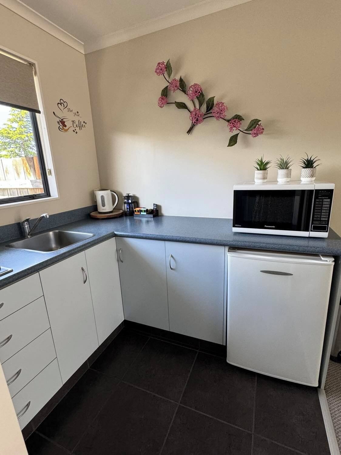 A compact kitchen with white cabinets, a sink, a kettle, a microwave, and a refrigerator. There is a decorative wall piece with pink flowers and a small window.
