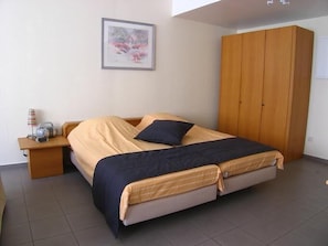 A bedroom with a bed, wooden wardrobe, bedside table with lamp, and a framed picture on the wall.