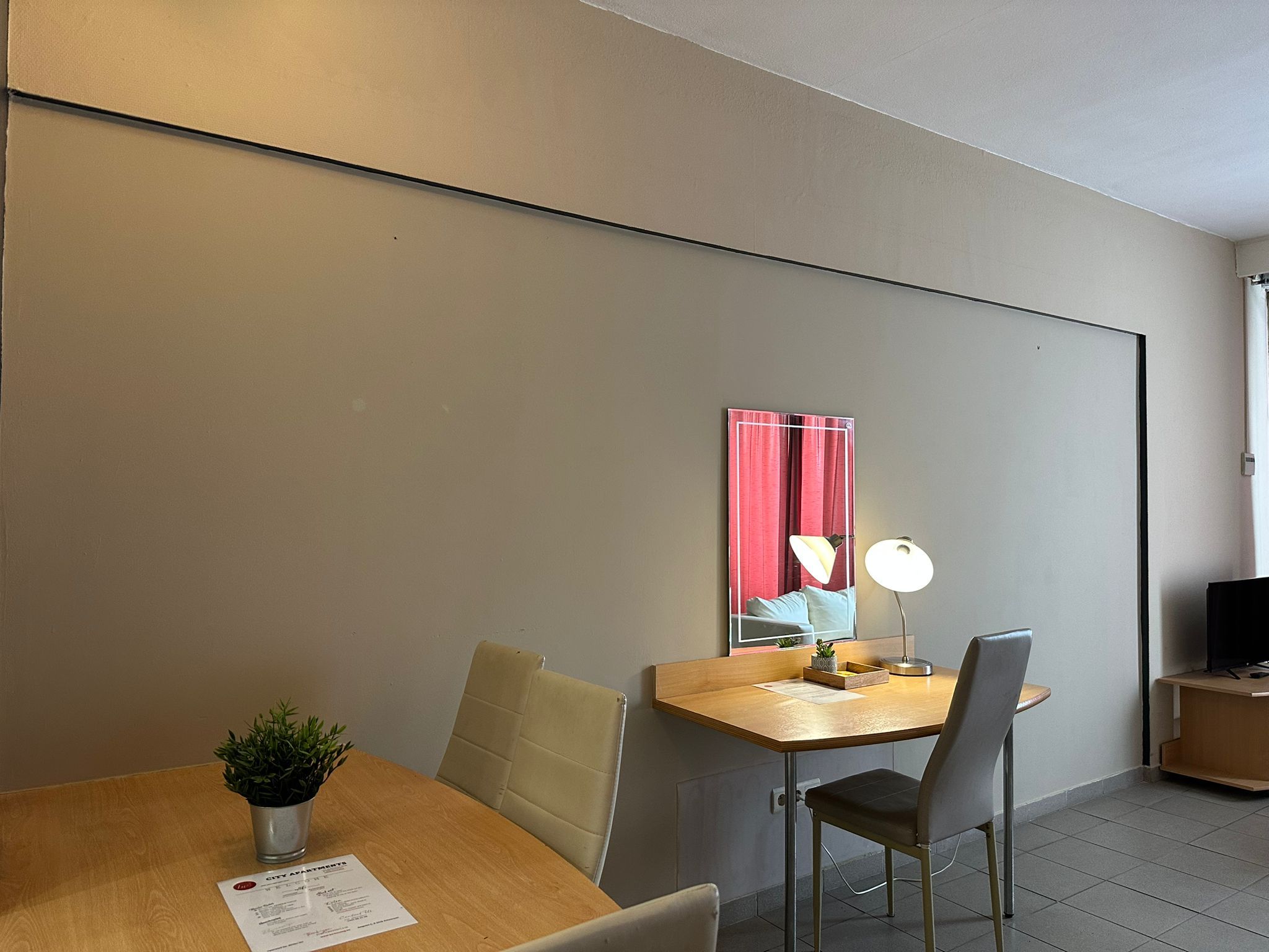 A modern room with a wooden desk, a chair, a small table with a plant, and a mirror with red curtains.